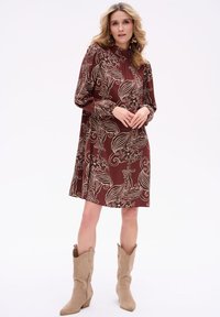 Brązowa sukienka z długimi rękawami, wysokim kołnierzem i kwiatowym wzorem w kolorze beżowym. Połączona z beżowymi kowbojkami.