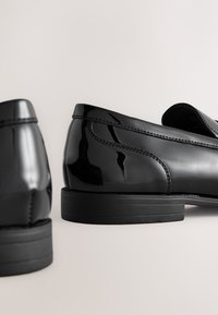 Sorte polerede loafers med en glat, blank overflade, buet design og en kontrasterende sort sål. Har syninger langs siderne.