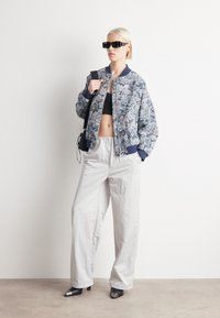 Veste bomber florale dans des tons de bleu avec des accents marine, associée à un pantalon large gris clair et des bottes à talons noires avec des bouts en argent.