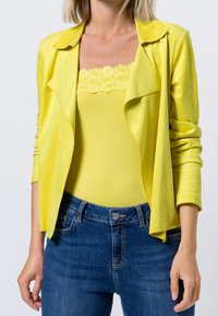 Żółta, długosleeve kurtka z miękkiego materiału z drapowaną przednią częścią, noszona na żółtej koronkowej tank topie i zestawiona z ciemnoniebieskimi dżinsami.