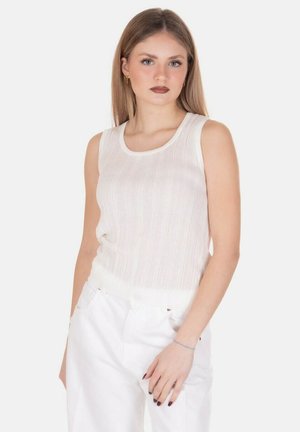 Giovane donna con capelli lisci castano chiaro che indossa un top bianco senza maniche e pantaloni bianchi, in piedi con una mano in tasca.