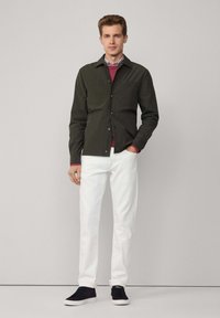 Ciemnozielona koszula zapinana na guziki z dwoma kieszeniami na piersi, nałożona na bordowy sweter, w połączeniu z białymi spodniami i czarnymi sneakersami.