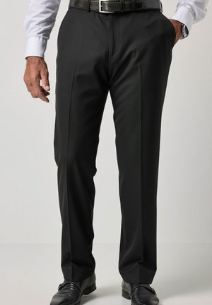 Mężczyzna ubrany w czarne eleganckie spodnie, czarny skórzany pasek, czarne eleganckie buty, białą koszulę, z jedną ręką w kieszeni i drugą swobodnie zwisającą.