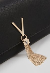 Pochette noire texturée avec un logo "V" doré prominent et un détail en frange composé de fines chaînes dorées.