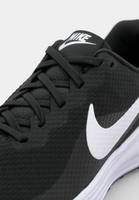 Black athletic shoe with textured mesh upper, grey laces, white swoosh logo, and white midsole. Close-up view highlights design details.