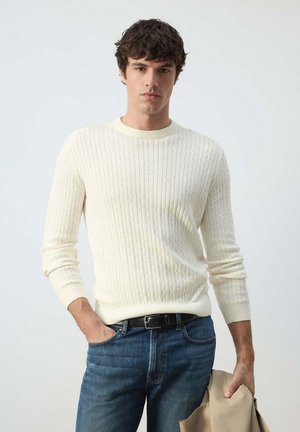 Man with curly dark hair wears cream cable-knit sweater and blue jeans, holding beige jacket in one hand, standing against plain background.