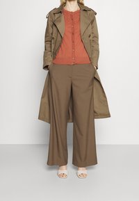 Hnědý trenčkot přes rusově červený top, široké hnědé kalhoty a béžové sandály. Výrazná textura na svetru a kabátu.