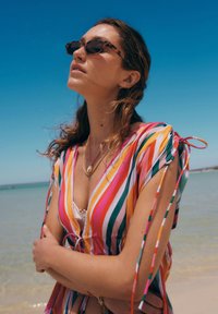 Copricostume da spiaggia a righe colorate in rosa, arancione e verde con maniche legate, indossato sopra una maglietta bianca, ambientato contro un cielo azzurro e acqua limpida.