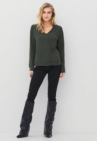 Olivine žalios spalvos ilgomis rankovėmis marškiniai su V formos iškirpte ir priekiu kišene, derinami su juodomis prisiderinančiomis kelnėmis ir juodais aukštais batais su sagtimis.