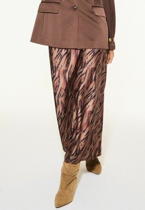 Brauner gemusterter Rock kombiniert mit brauner Jacke und beige Wildleder-Stiefeletten mit spitzer Zehenpartie vor hellem Hintergrund.