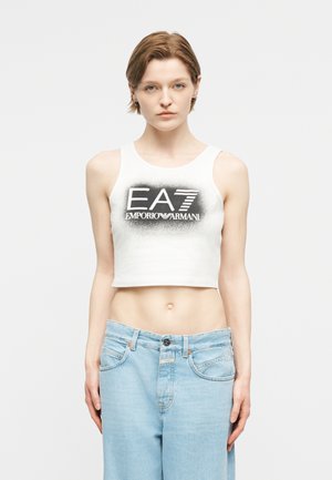Νέος άνθρωπος που φοράει λευκό κοντό τοπ με μαύρο λογότυπο "EA7 Emporio Armani" και ανοιχτό μπλε τζιν, στέκεται μπροστά σε απλό φόντο.