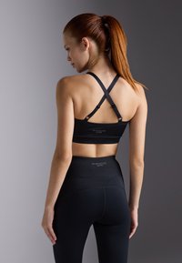 Crna sportska bralette sa leđnim trakama u obliku križa. Visoki crni tajice s teksturiranim pojasom. Logotip branda vidljiv na oba komada odjeće.
