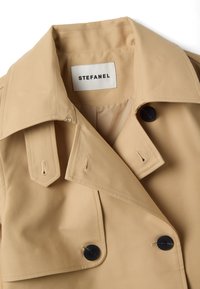 Beiger Trenchcoat mit weitem Kragen, schwarzen Knöpfen und einem weißen Etikett mit der Aufschrift "STEFANEL" am Halsansatz.