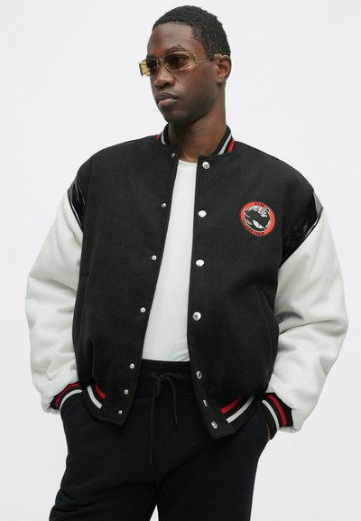 Black and white varsity jacket with red accents, metallic buttons, and an embroidered logo. Features a wool body and leather sleeves.