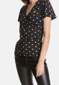 Svart topp med prickar, korta volangärmar, omlott-design och en liten logotyp på vänster sida, tillverkad av texturerad tyg. Kombination med svarta läderbyxor.