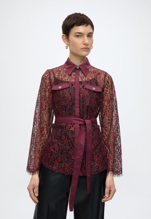 Femme aux cheveux courts et foncés porte un chemisier en dentelle florale bordeaux avec col en satin, poches poitrine et ceinture assortie à la taille, porté avec un pantalon noir.