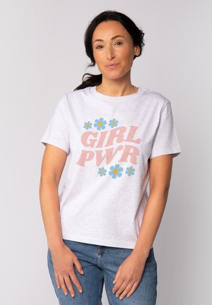 Frau mit dunklem Haar, die ein weißes T-Shirt mit dem Schriftzug "GIRL PWR" und blauen Blumen trägt, kombiniert mit blauer Jeans, steht vor einem einfarbigen Hintergrund.
