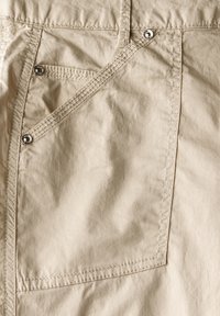 Pantaloni in cotone beige con patta della tasca diagonale, minuteria in metallo e dettagli di cucitura visibili su un tessuto testurizzato.