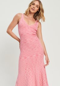 Ružová maxi šaty bez rukávov s výstrihom do V a textúrovaným, pruhovaným vzorom látky. Silueta je na vrchu priliehavá a smerom dole sa rozširuje.