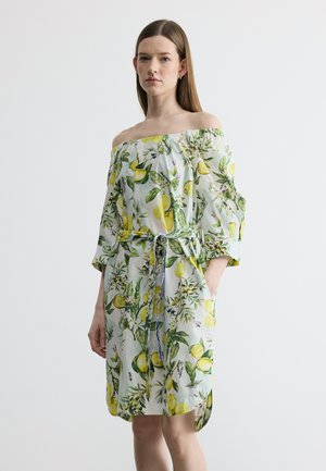 Woman wearing off-shoulder white dress with lemon and green leaf print, hands in pockets, standing against a plain light background.