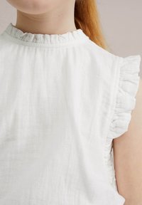 White sleeveless top with a ruffled neck and armhole trim. Made from lightweight fabric, featuring a soft, textured appearance.