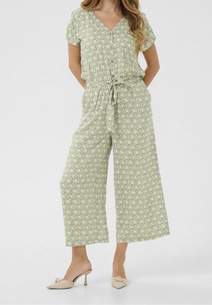 Combinaison à motifs verts avec encolure en V, manches courtes, taille ajustable par cordon et coupe pattes d'éléphant, associée à des mules à talons beiges.