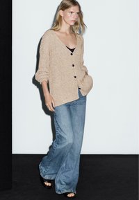 Ljusbeige stickad cardigan med V-ringning, knäppning och uppvikta ärmar, kombinerad med ljusblå vida jeans och svarta sandaler med öppna tår.
