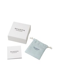 Vit kvadratisk låda med "SKAGEN DANMARK" tryckt i svart, tillsammans med en ljusblå tygpåse och en liten broschyr med samma logotyp.