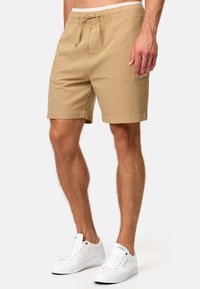Pantalones cortos de algodón color tostado con cintura ajustable, que cuentan con bolsillos laterales, textura suave y un corte relajado. Combinados con zapatillas blancas.