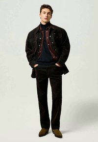 Tmavohnedá kordurová bunda cez vínovú košeľu a čierny rolák, doplnená o zladené nohavice a hnedé semišové topánky.