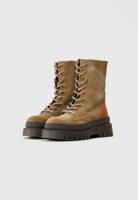 Botas de ante marrón con cordones, suela negra gruesa y un acento de cuero marrón en el talón. Presentan ojales de metal y una superficie texturizada.