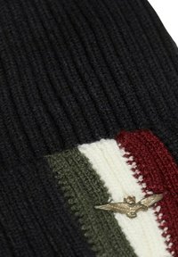 Beanie lavorato a maglia di colore nero, con texture a righe. Accentuato da strisce crema, verde e rosso. Include un'emblema metallico di un'aquila.