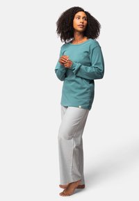 Turquoise lange mouw top met ribbelstructuur, gecombineerd met lichtgrijze gestreepte pyjamabroek. Opvallend merketiket op de broek. Voeten zijn bloot.