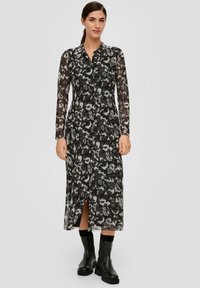 Schwarz-weiß gemustertes Kleid mit Kragen, langen Ärmeln, durchgehender Knopfleiste und einem Schlitz am unteren Ende. Getragen mit schwarzen Stiefeletten.