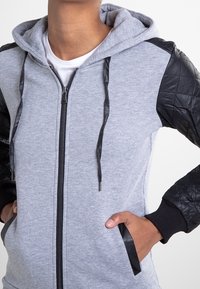 Personne portant un sweat à capuche gris zippé avec des manches en cuir matelassé noir et une bordure noire sur les poches et les cordons, mains dans les poches.