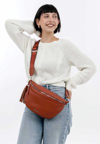 Borsa a tracolla in pelle marrone con chiusura a zip, forma arrotondata e dettaglio con nappina. Indossata con un maglione di lana color crema e jeans.