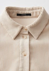 Camicia beige a coste con colletto classico, dotata di tre bottoni rotondi e un'etichetta che recita "un giorno." Texture liscia con righe verticali.