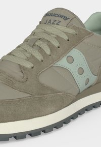 Κοντινή λήψη ενός μπεζ και ανοιχτοπράσινου sneaker Saucony Jazz, που δείχνει υφασμένα κορδόνια, σουέντ και υφασμάτινα υλικά, καθώς και σόλα με μοτίβο για καλύτερη πρόσφυση.