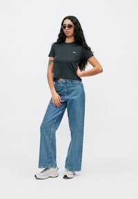 Mørkegrå cropped t-skjorte med lite logo; lyseblå, løst sittende jeans; hvite sneakers. Personen har runde solbriller og langt hår.