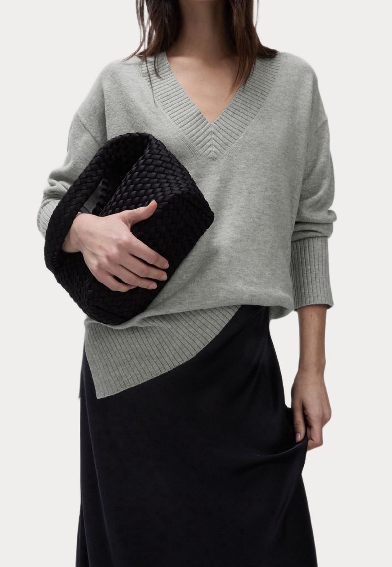 Femme portant un pull gris clair à col en V et une jupe noire, tenant un sac à main noir tressé d'une main.