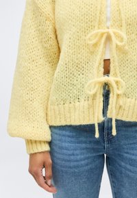 Κοντινό πλάνο ενός ατόμου που φοράει ένα απαλό κίτρινο πλεκτό ζακέτα, δεμένο με δύο φιόγκους πάνω από λευκή μπλούζα και μπλε τζιν.
