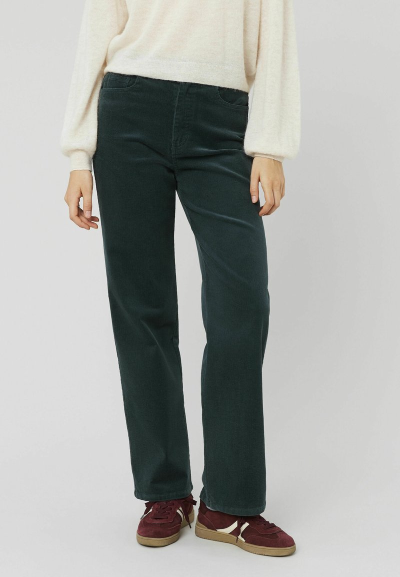 Pantalon en velours côtelé vert avec une coupe droite, doté de poches latérales et d'un tissu texturé. Associé à des baskets bordeaux.