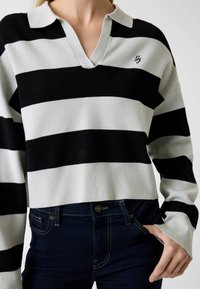 Pull rayé noir et blanc avec un col à revers et un ourlet côtelé. Associé à un jean bleu foncé, avec une fermeture à boutons et une poche.