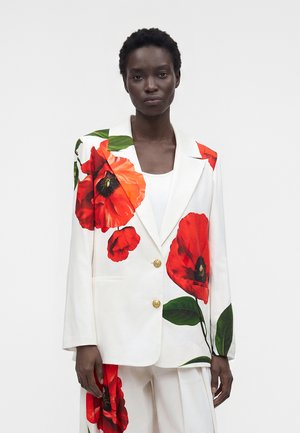 Femme portant un blazer blanc et un pantalon avec de grandes imprimés de fleurs de coquelicot rouges, debout devant un fond blanc uni.