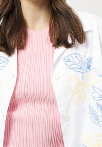Camicia bianca ricamata con motivi floreali blu e gialli, indossata su un top a coste rosa chiaro. Bottoni visibili sulla camicia.