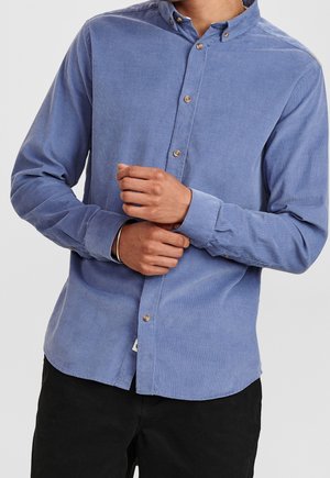 Homme portant une chemise en velours côtelé bleue à boutons, ajustant son poignet, avec un pantalon noir visible sous la chemise.