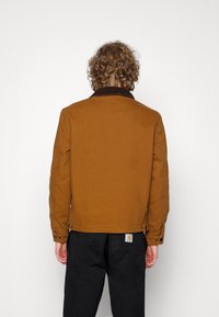 Brun lærredjakke med en corduroy krave. Har en lige pasform, knapmanchetter og en ryglabel. Bæres med sorte bukser.