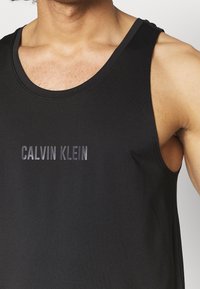Czarny top na ramiączkach wykonany z gładkiego materiału, z dekoltem w serek i nadrukowanym logo "CALVIN KLEIN" w ciemnoszarym kolorze na klatce piersiowej.