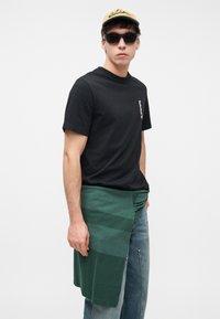 Νέος άνδρας που φοράει μαύρο t-shirt Timberland, γυαλιά ηλίου, μπεζ καπέλο, πράσινο πουλόβερ δεμένο γύρω από τη μέση και ανοιχτό μπλε τζιν, στέκεται μπροστά σε λευκό φόντο.