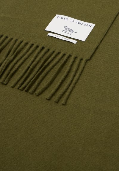 Pătura din lână verde măsliniu cu margini franjurate, având o etichetă albă care citește "TIGER OF SWEDEN" și "MADE IN ITALY." Textură moale.